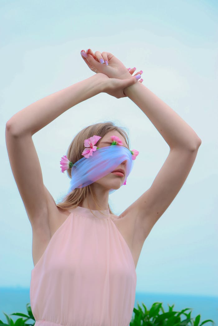 A woman in a pastel dress poses with a floral blindfold, creating a serene and artistic image by the sea.