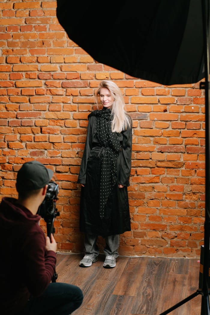 Fashion model in black dress posing against brick wall in studio setting.